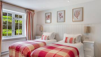 A bedroom with two beds, a window with curtains, and three framed botanical prints on the wall.