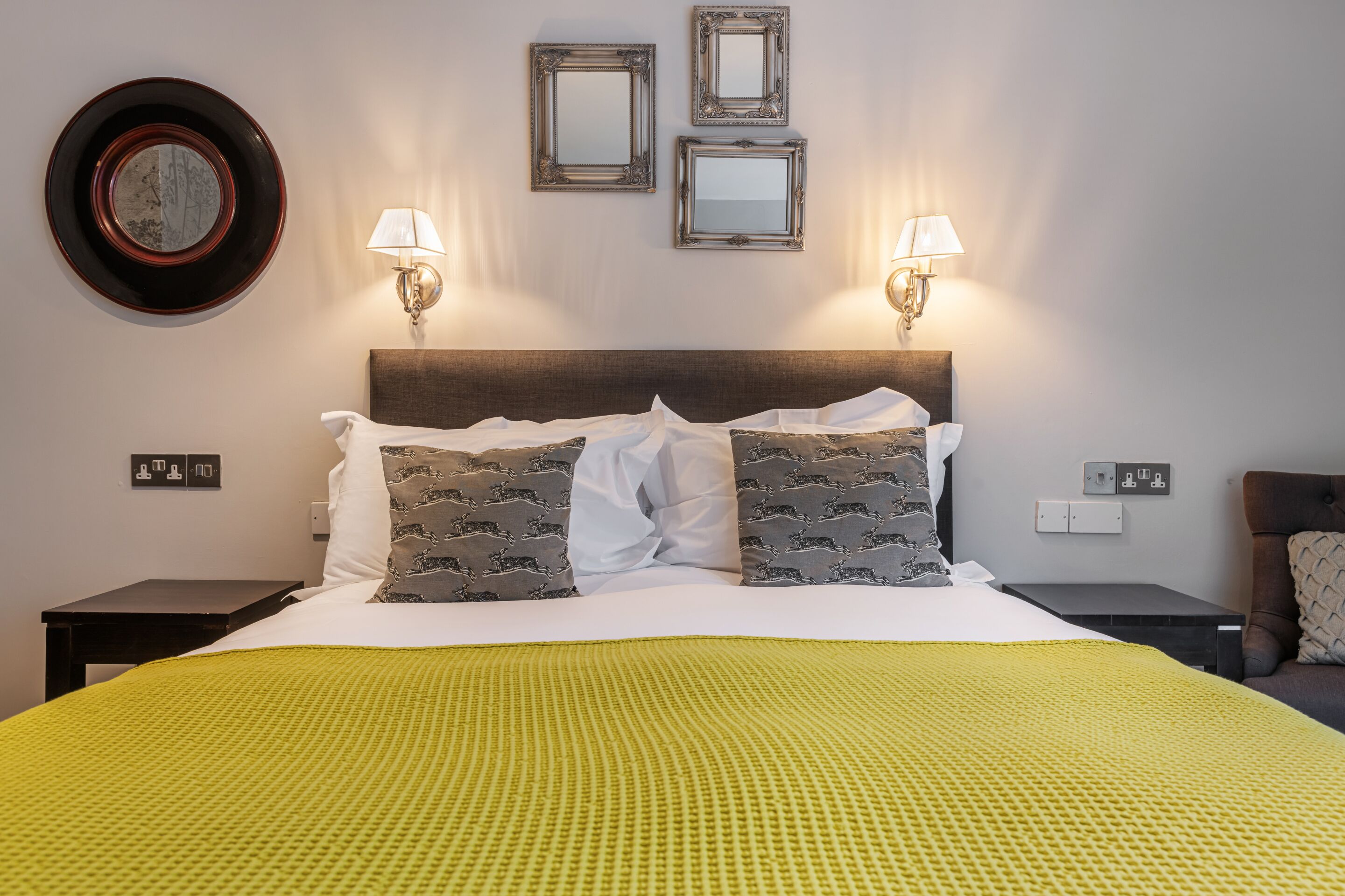 A neatly made bed with a yellow blanket, two wall-mounted lamps, and three framed mirrors on the wall.