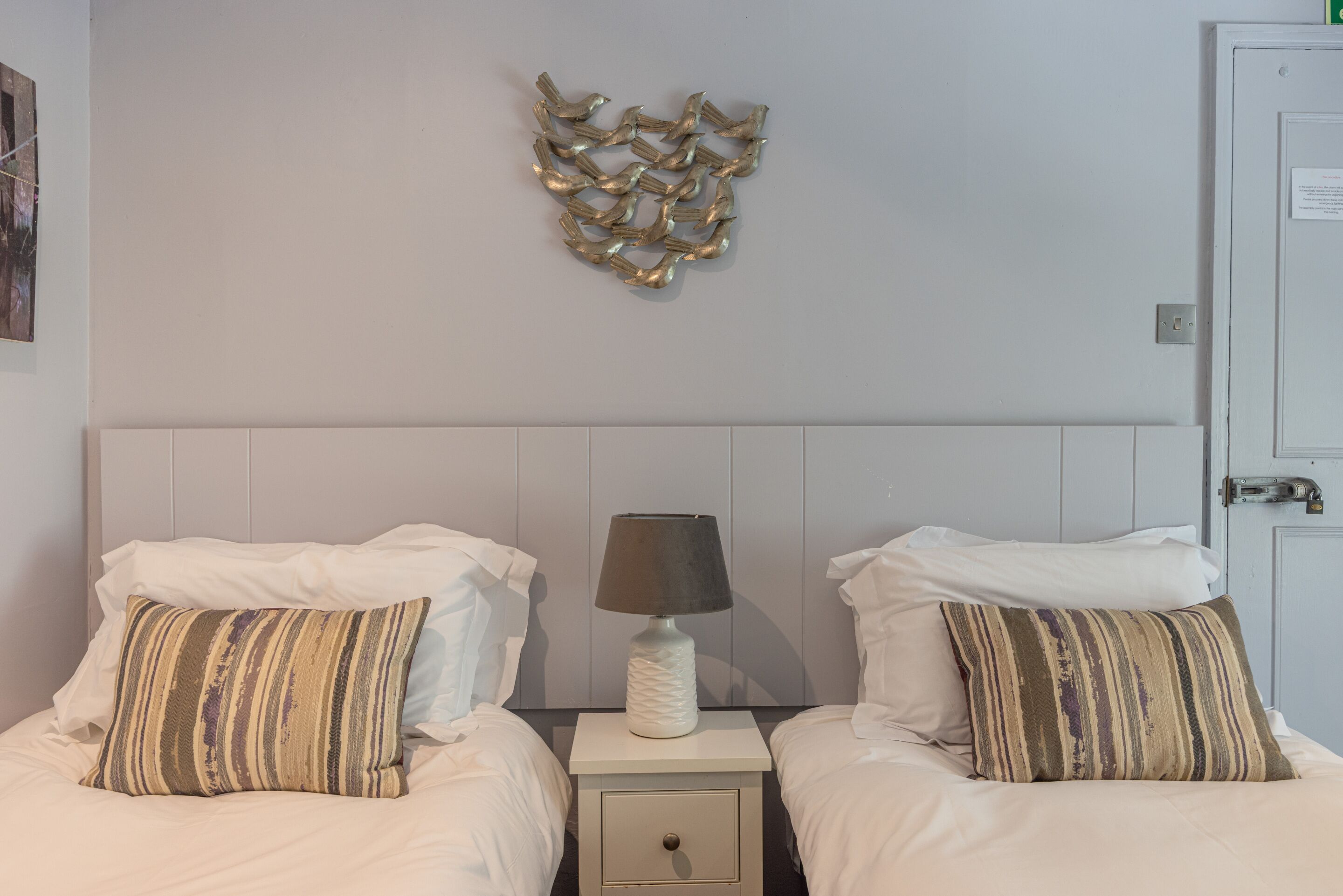 A hotel room with two beds, each with striped pillows, a bedside table with a lamp, and a decorative wall piece.