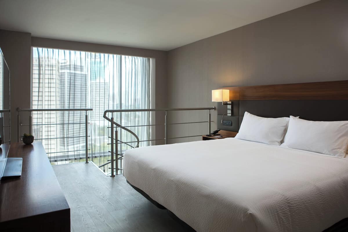 A hotel room with a large bed, a desk, and a view of the city through large windows.