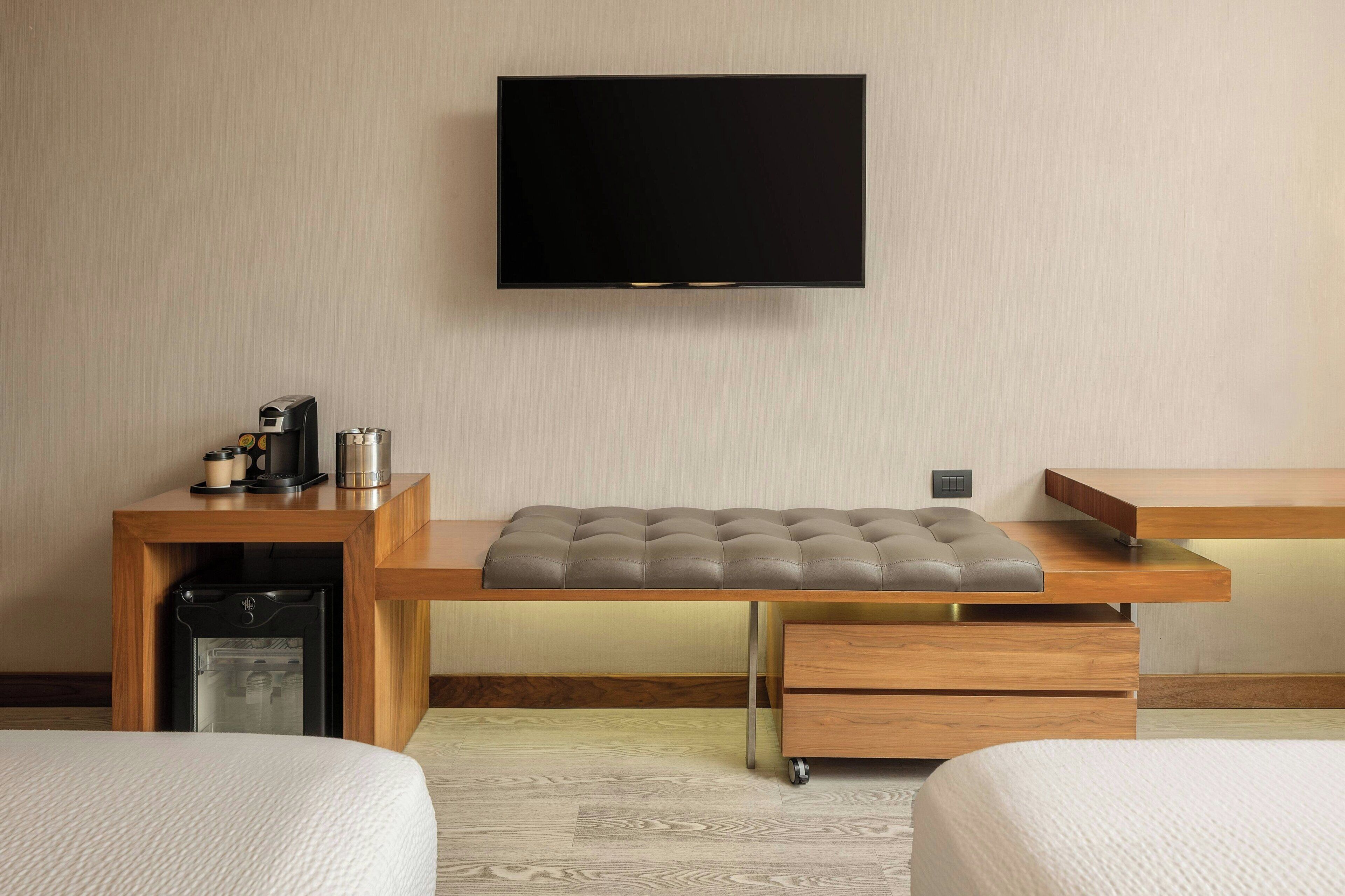 A hotel room with a bed, a desk with a flat-screen TV, a coffee machine, and a mini-fridge.
