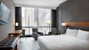 A hotel room with a large bed, a desk, a bench, two armchairs, a small table, and a TV mounted on the wall.
