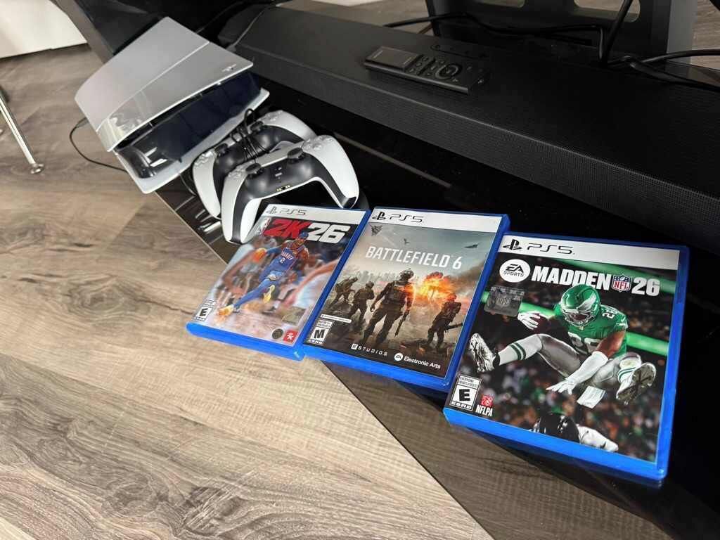 A gaming setup with a PlayStation 5 console, a VR headset, and three game cases: Battlefield 6, Madden NFL 26, and a third game with a blue cover.