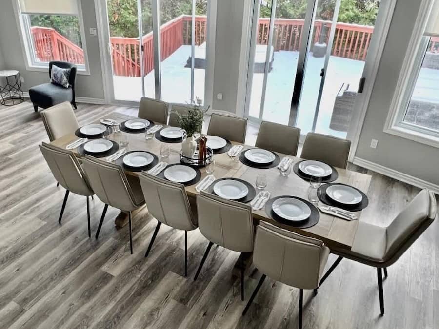 A dining area with a large table set for 10, surrounded by chairs, with a view of a red deck through glass doors.