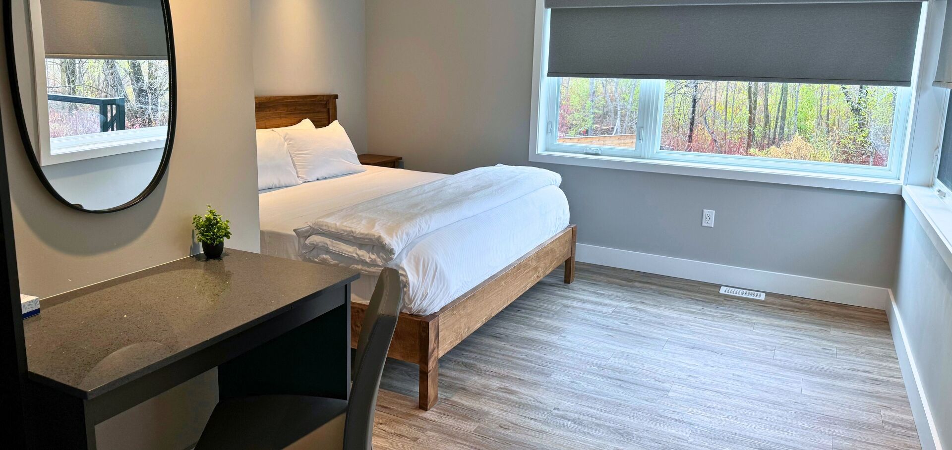 A bedroom with a bed, a desk, a mirror, and a window with a view of trees.