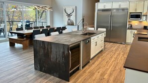 A modern kitchen with a large island, stainless steel appliances, and a dining area with a view of the outdoors. - Orchard Run Chalet (The Blue Mountains)