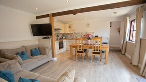 A modern living room with a sofa, a dining table with chairs, a kitchen area, and a TV mounted on the wall. - Newclose Farm (Yarmouth)