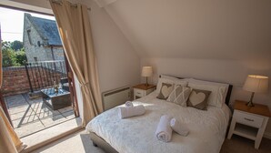 A bedroom with a large bed, two bedside tables with lamps, a view of a stone building through a balcony door, and a small outdoor seating area. - Newclose Farm (Yarmouth)