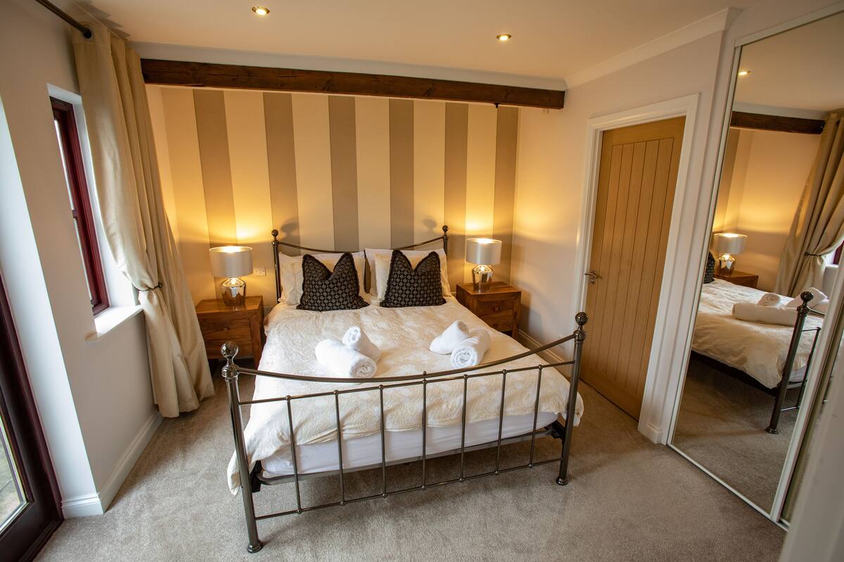 A neatly made bed with white linens and two pillows, a bedside table with a lamp, a window with curtains, and a mirror reflecting the room.