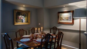 A dining room with a wooden table set for six, surrounded by six chairs. There are two framed paintings on the walls, a floor lamp, and candles on the table.