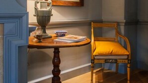 A corner room with a wooden chair, a table with a lamp, and a framed picture on the wall.