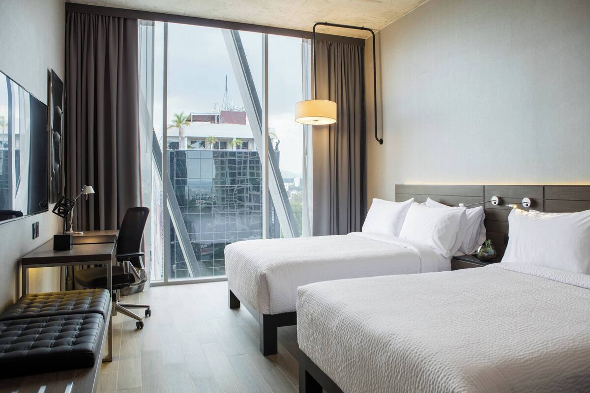 Habitación de hotel con dos camas, un escritorio, una silla y un ventanal con vistas a edificios.