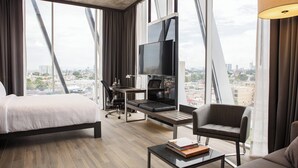 Una habitación de hotel moderna con una cama, un escritorio con computadora, televisor, sofá y vista a la ciudad.