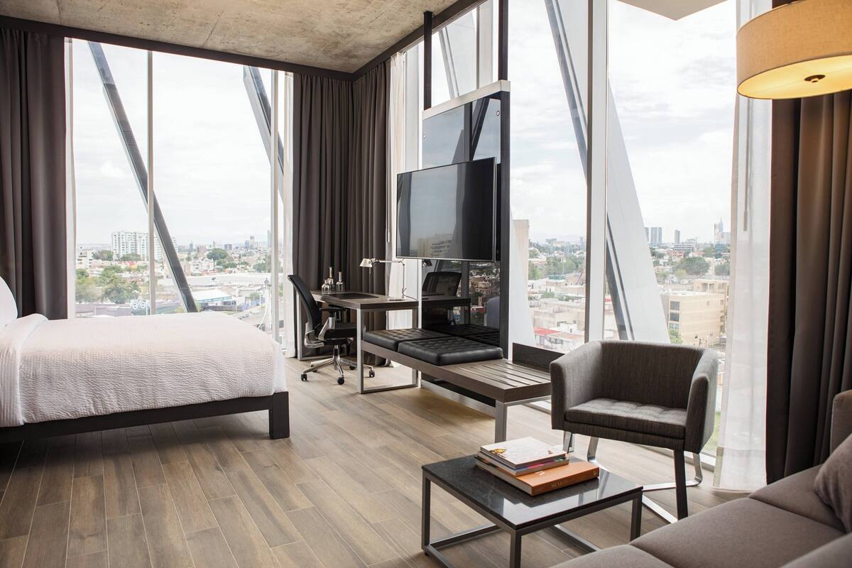 Una habitación de hotel moderna con una cama, un escritorio con computadora, televisor, sofá y vista a la ciudad.