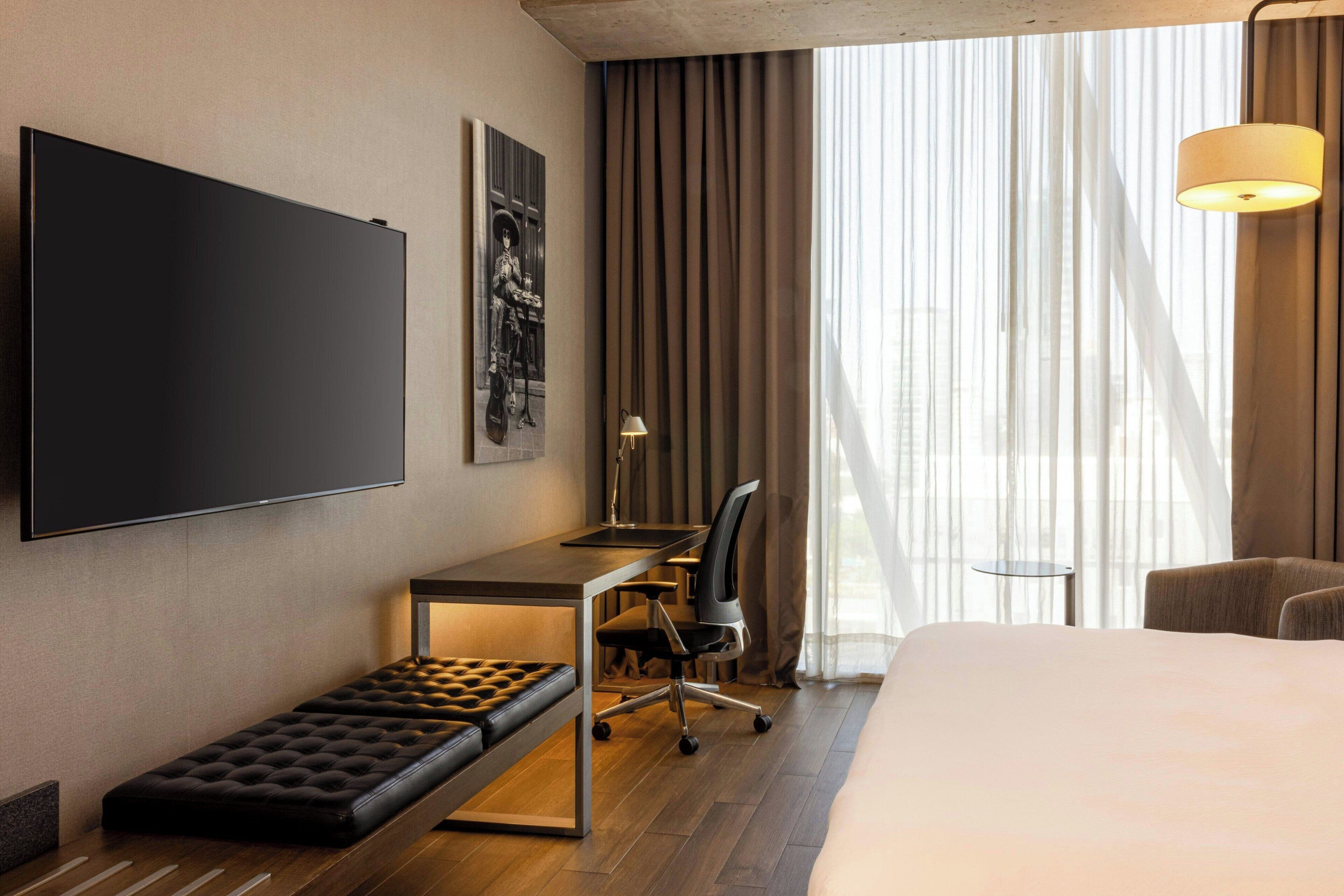 A hotel room with a bed, a desk with a chair, a bench, a TV mounted on the wall, and a window with curtains.