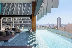 Ein Rooftop-Infinity-Pool mit Liegestühlen, einer Holzterrasse und Blick auf die Stadt.