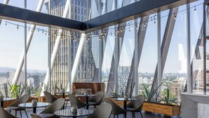 Un área de comedor moderna en la azotea con mesas y sillas, ventanas amplias y vistas a la ciudad.