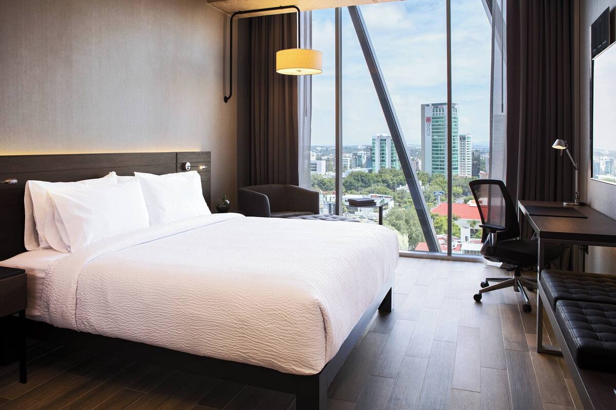 Habitación de hotel moderna con una cama grande, un escritorio y vistas de la ciudad a través de amplias ventanas.