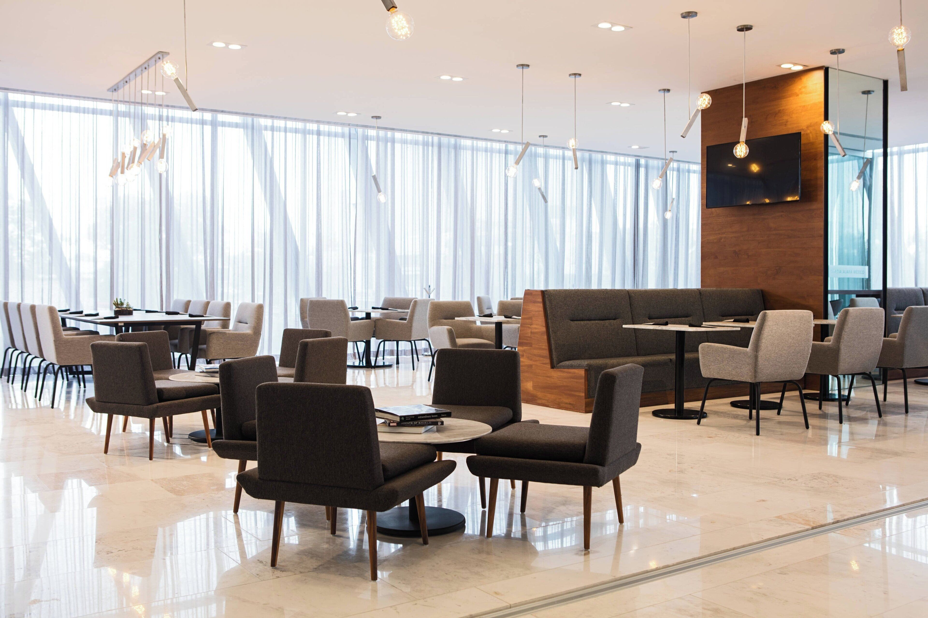 A modern, well-lit cafe with a variety of seating options including tables and sofas, large windows with sheer curtains, and a flat-screen TV mounted on a wooden panel.