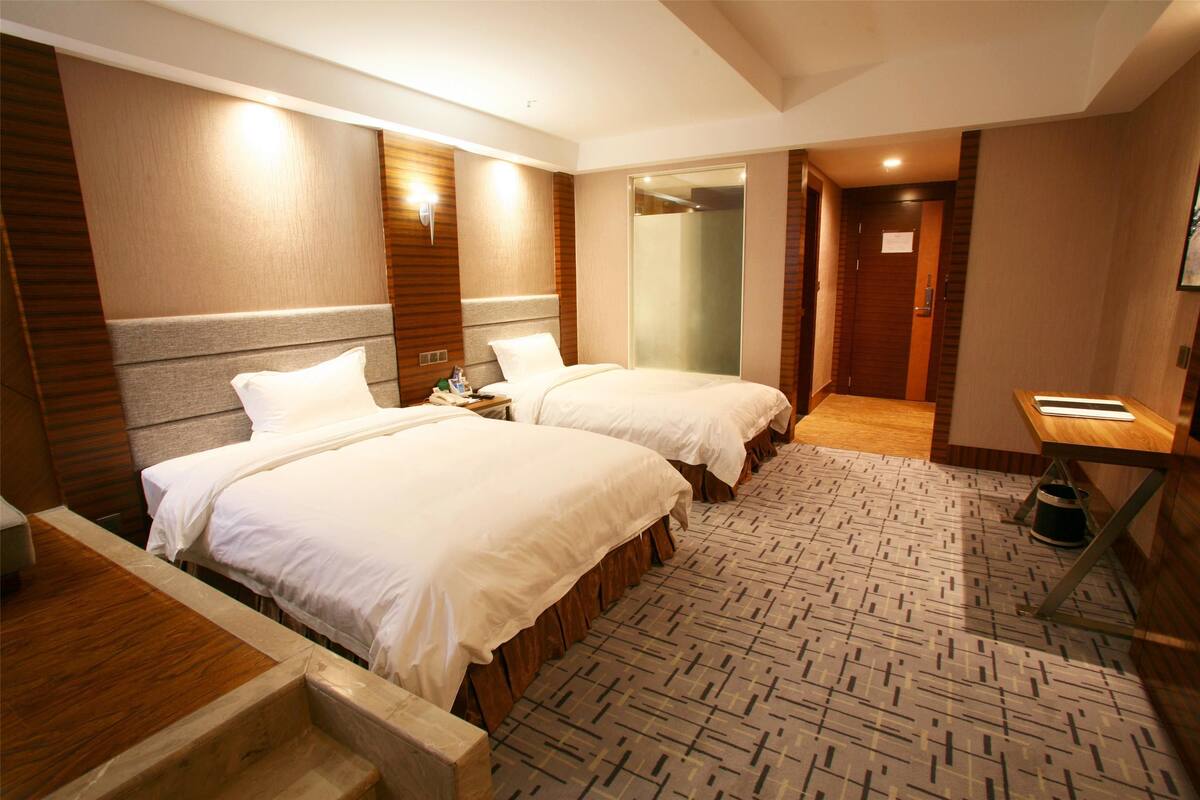 A hotel room with two beds, a wooden headboard, a small desk, and a bathroom with a shower.