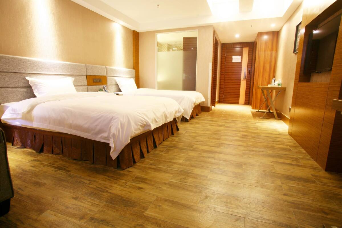 A hotel room with two beds, a wooden floor, a TV mounted on the wall, and a small table with a bottle.