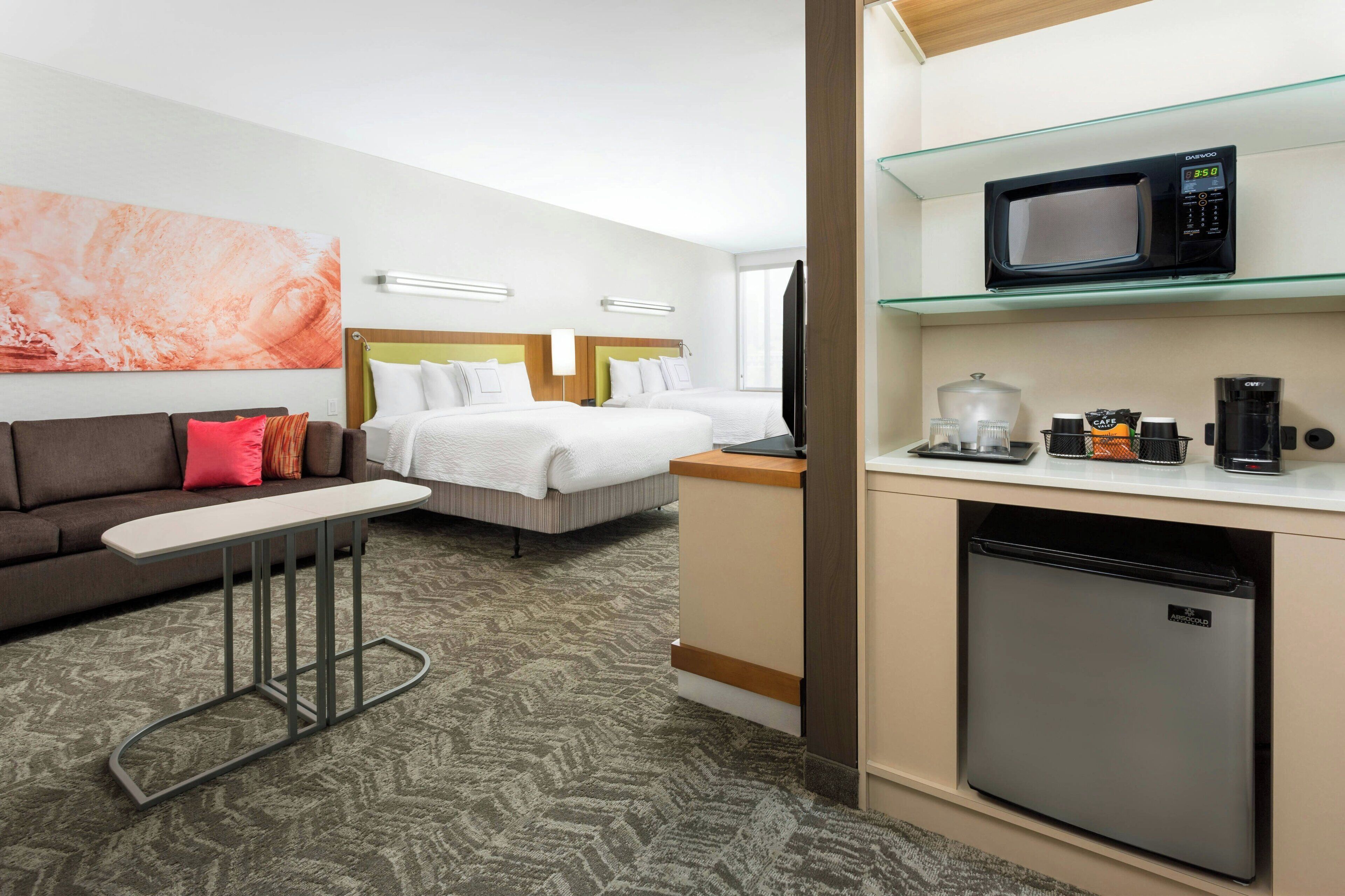 A hotel room with a bed, a sofa, a small table, a microwave, a coffee maker, and a mini fridge.