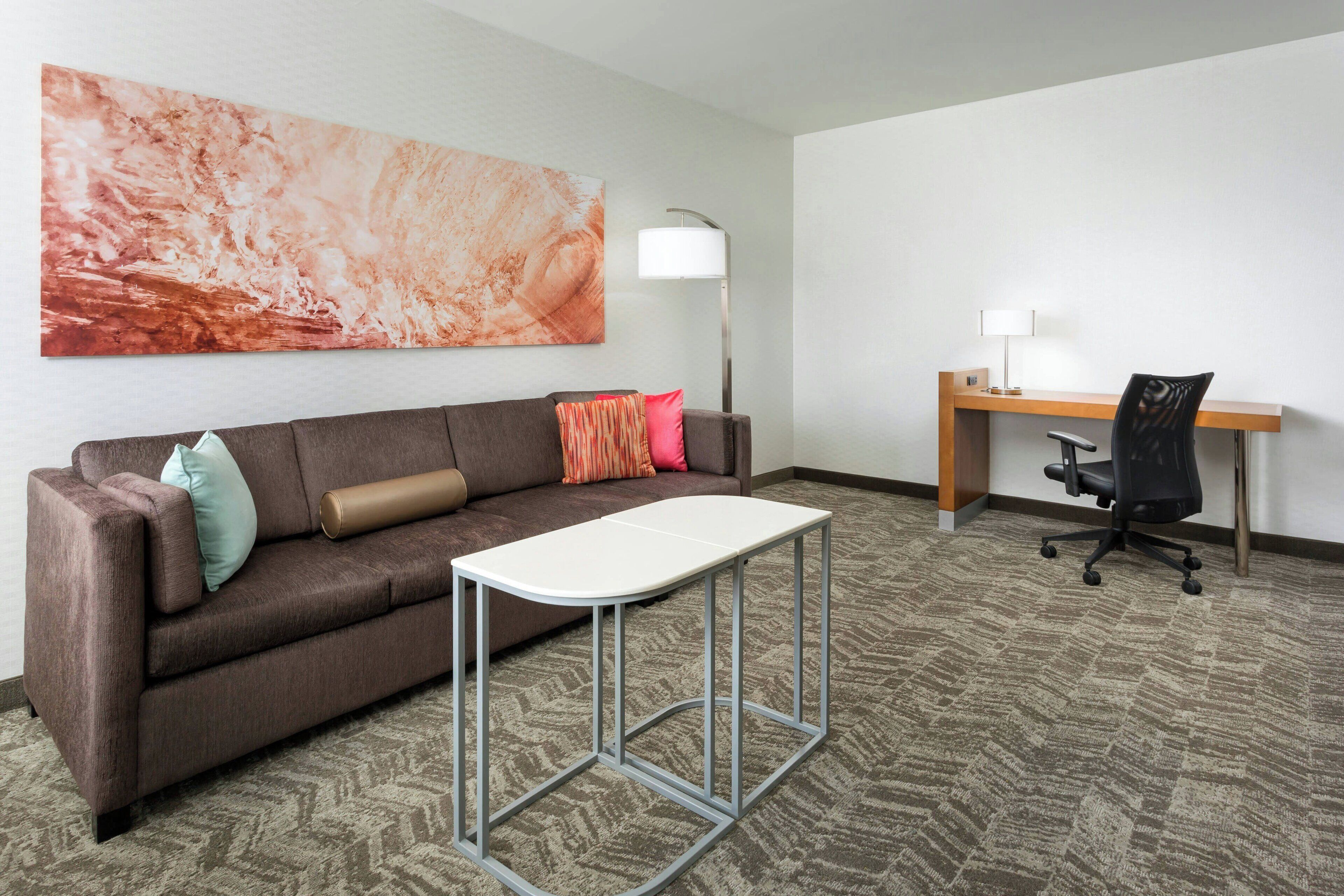 A hotel room with a brown sofa, a white table, a desk with a chair, a floor lamp, and a large abstract painting on the wall.