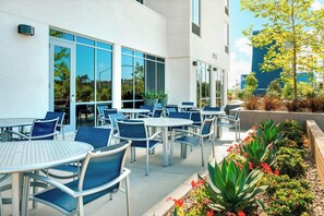 An outdoor seating area with tables and chairs, a building with large windows, and a landscaped garden with plants and flowers. - Springhill Suites San Diego Mission Valley (San Diego)
