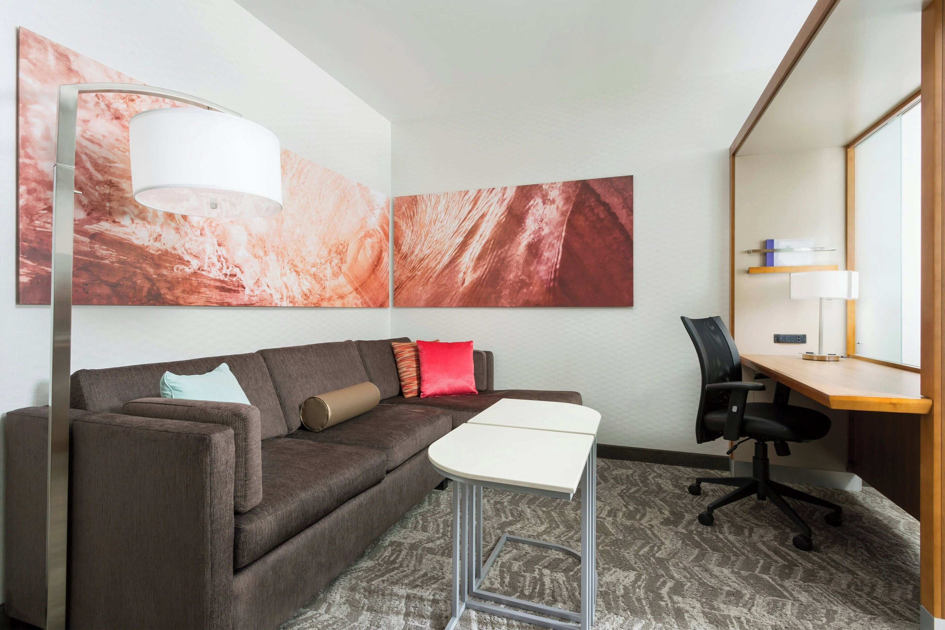 Habitación de hotel moderna con sofá, una pequeña mesa, escritorio y una silla. Hay pinturas abstractas en la pared y una lámpara en el suelo.