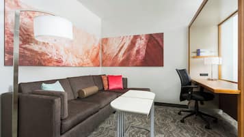 Habitación de hotel moderna con sofá, una pequeña mesa, escritorio y una silla. Hay pinturas abstractas en la pared y una lámpara en el suelo.