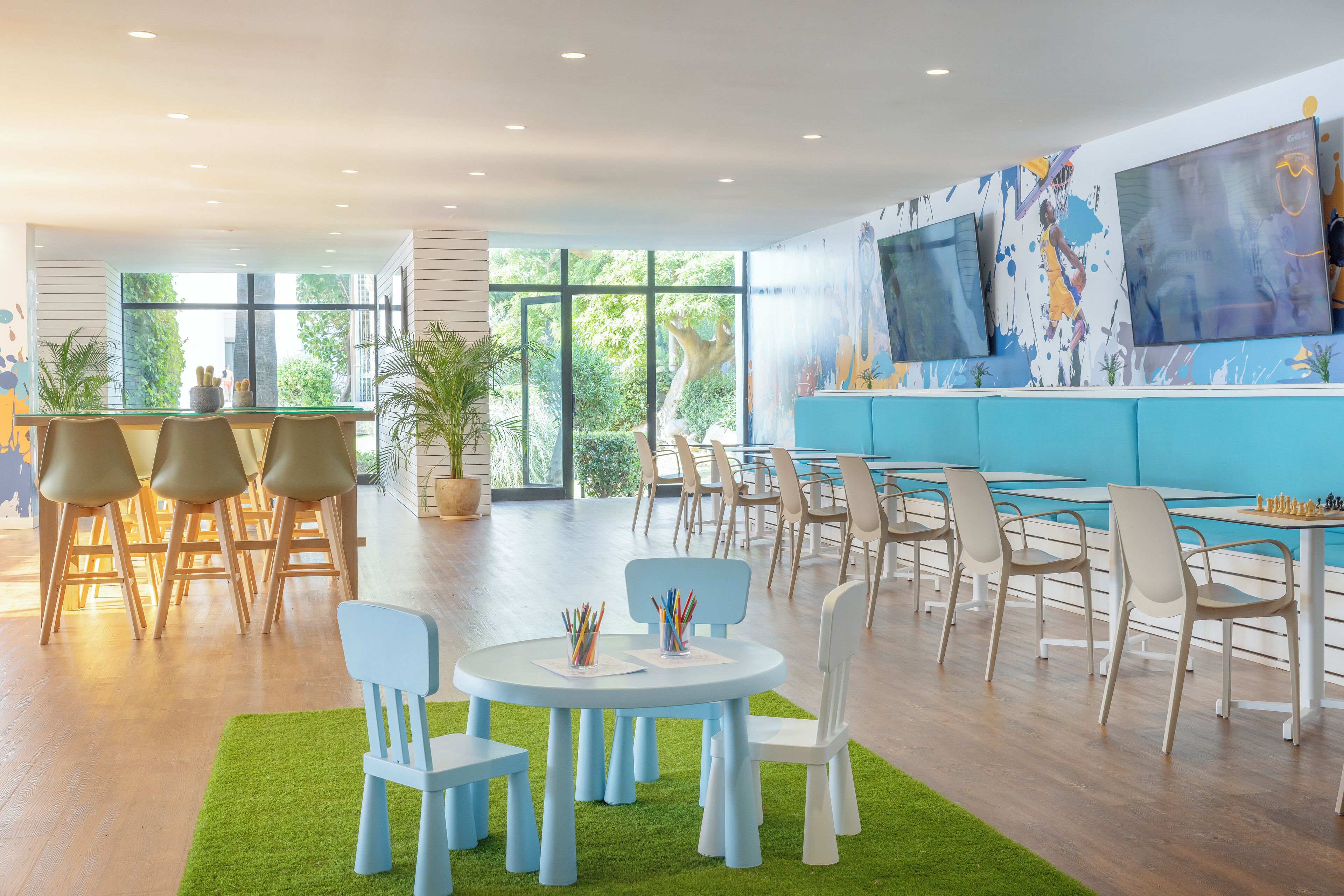 A modern indoor space with multiple seating areas, including round tables and chairs, and a large TV screen on the wall.