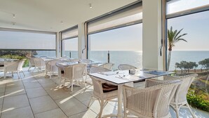 A dining area with white wicker furniture and large windows overlooking the sea.