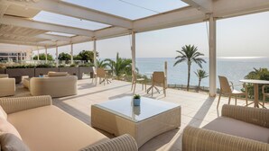A covered outdoor seating area with wicker furniture, a glass coffee table, and a view of the sea.