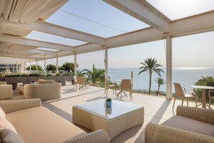 Un espacio de estar al aire libre techado con mobiliario de mimbre, una mesa de centro de vidrio y vistas al mar.