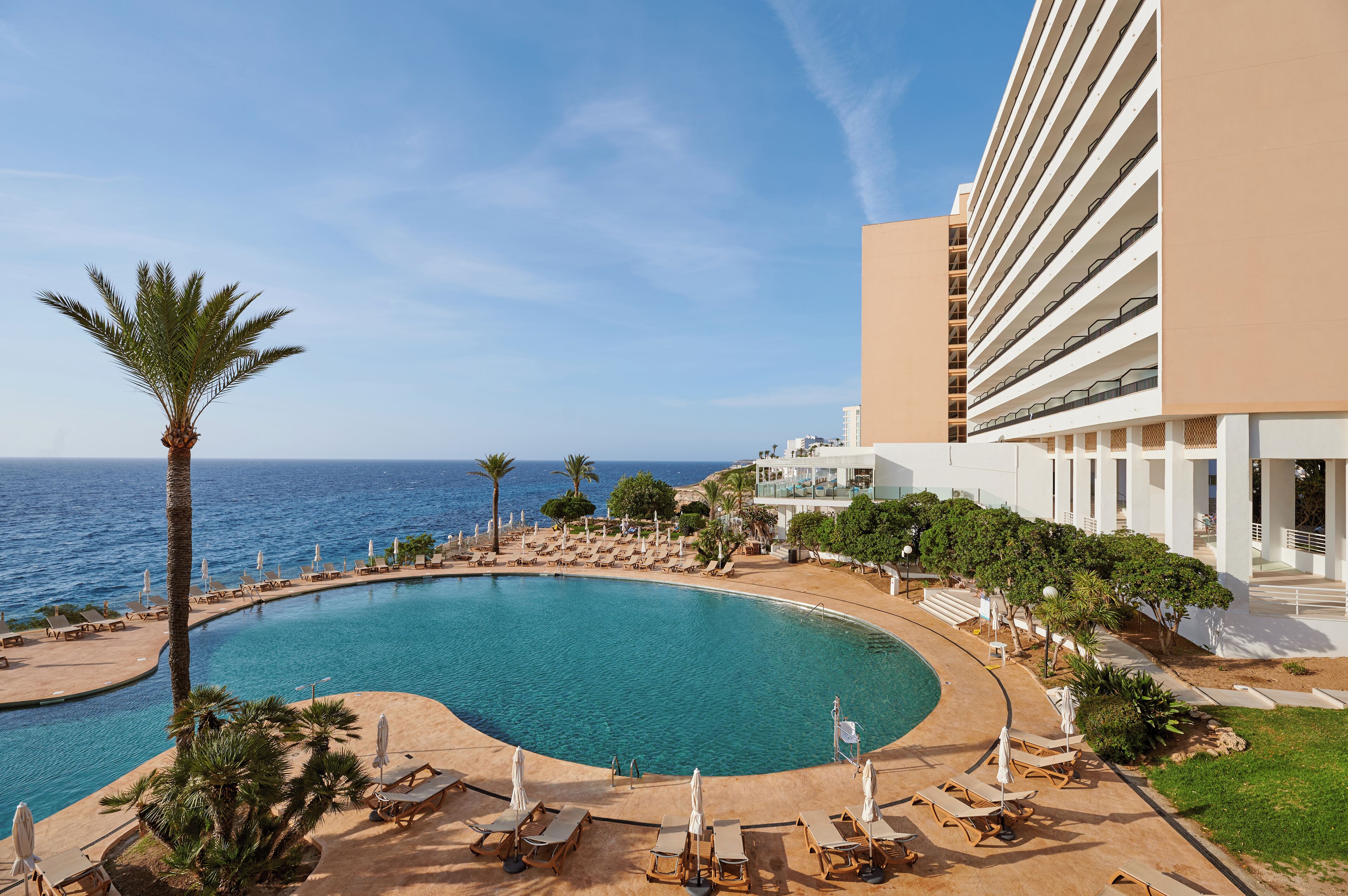En pool på ett semesterhotell med en böjd design, omgiven av solstolar och parasoller, och med utsikt över havet.