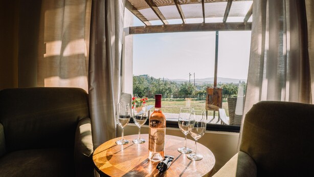 Una mesa con una botella de vino y copas, dos sillas y vistas a un paisaje a través de una ventana.