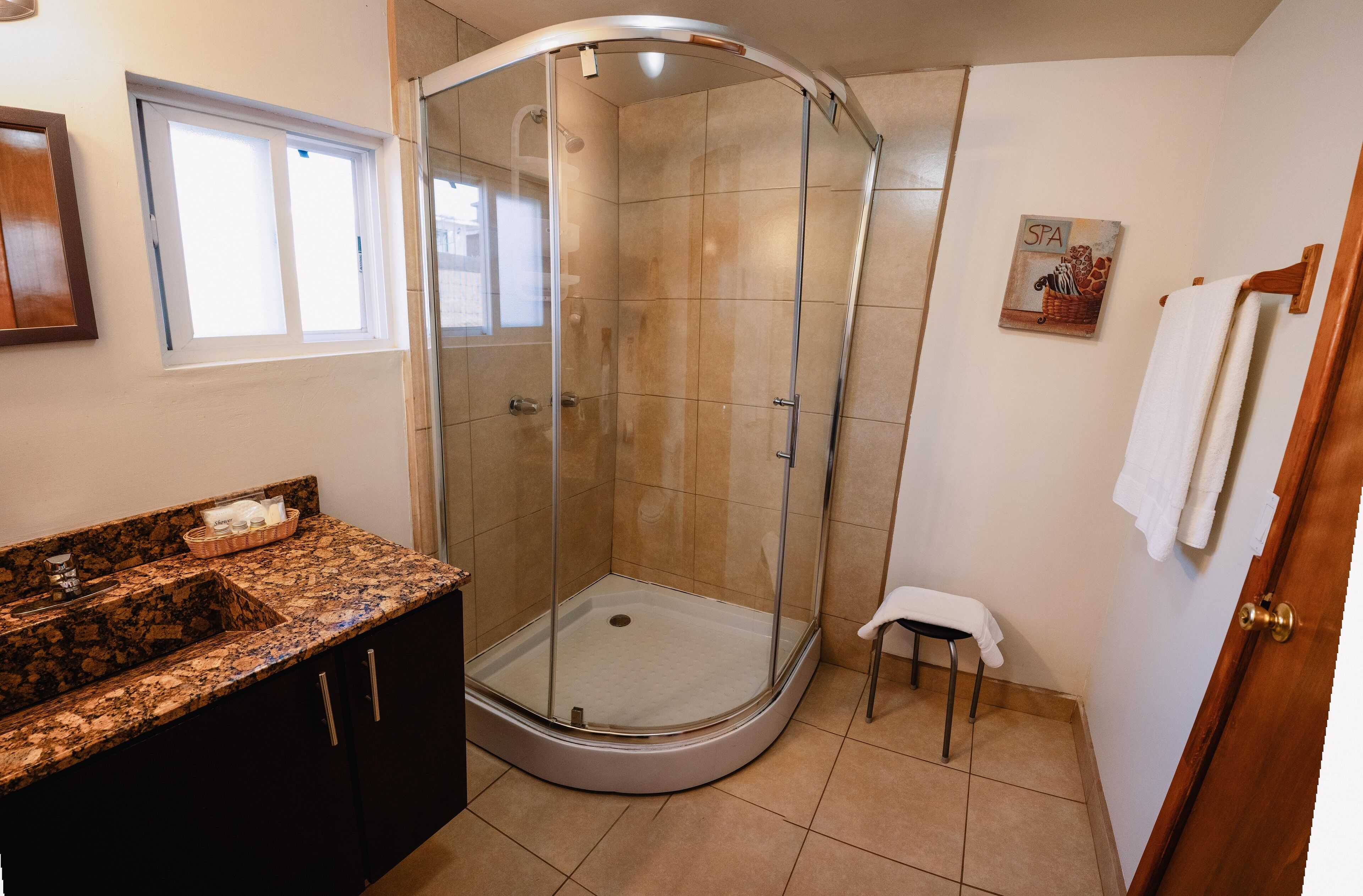 Un baño con ducha envuelta en vidrio, lavamanos con encimera de granito y un taburete pequeño.