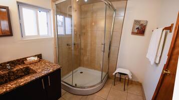 Un baño con ducha envuelta en vidrio, lavamanos con encimera de granito y un taburete pequeño.