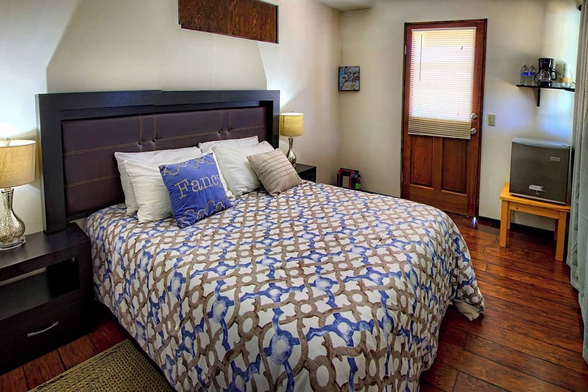 A bedroom with a large bed, two bedside tables with lamps, a wooden headboard, a door with a window, a small table with a TV, and a shelf with items.