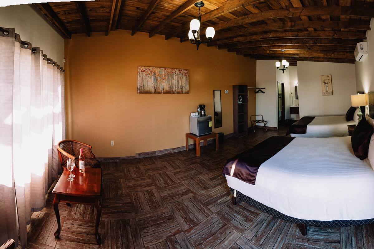 A hotel room with a large bed, a wooden floor, a desk with a chair, a small table with a coffee machine, and a mirror on the wall.