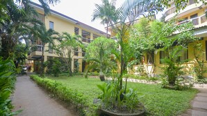 Un patio con un sendero, rodeado de abundante vegetación y un edificio amarillo con balcones.