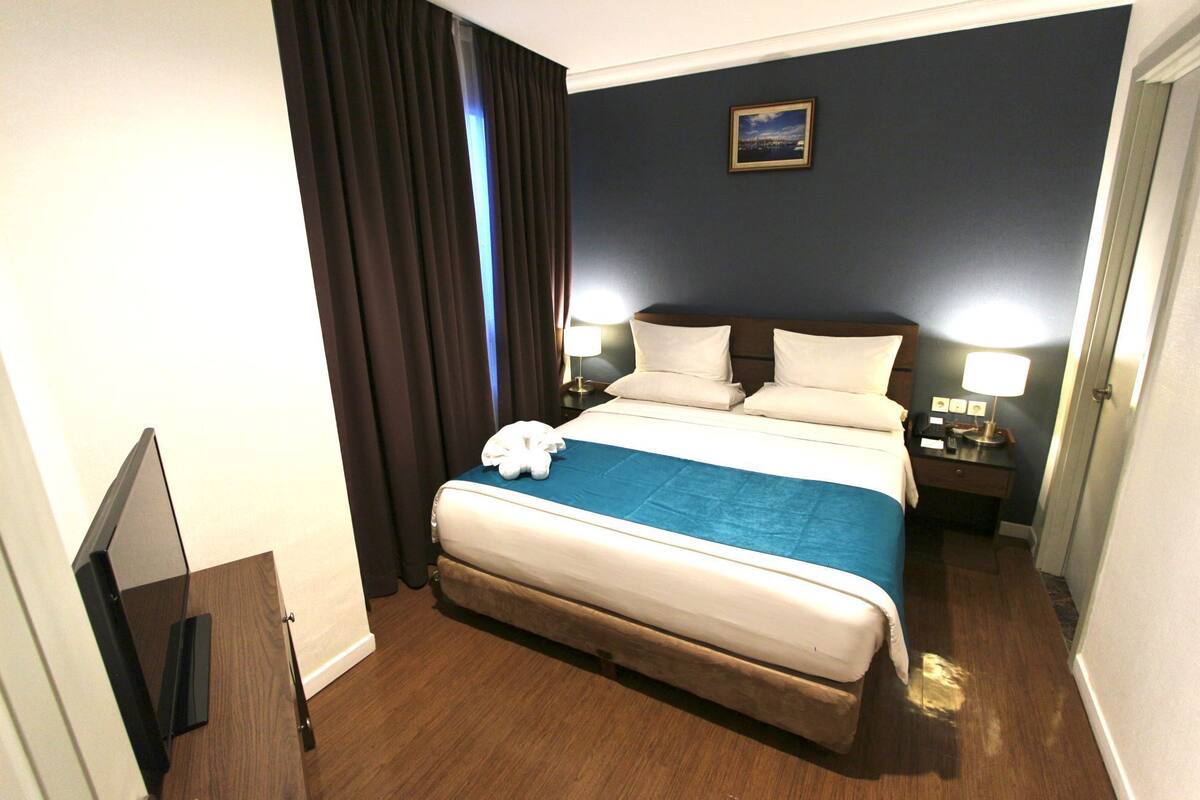 A hotel room with a large bed, dark walls, a framed picture, and a desk with a lamp.