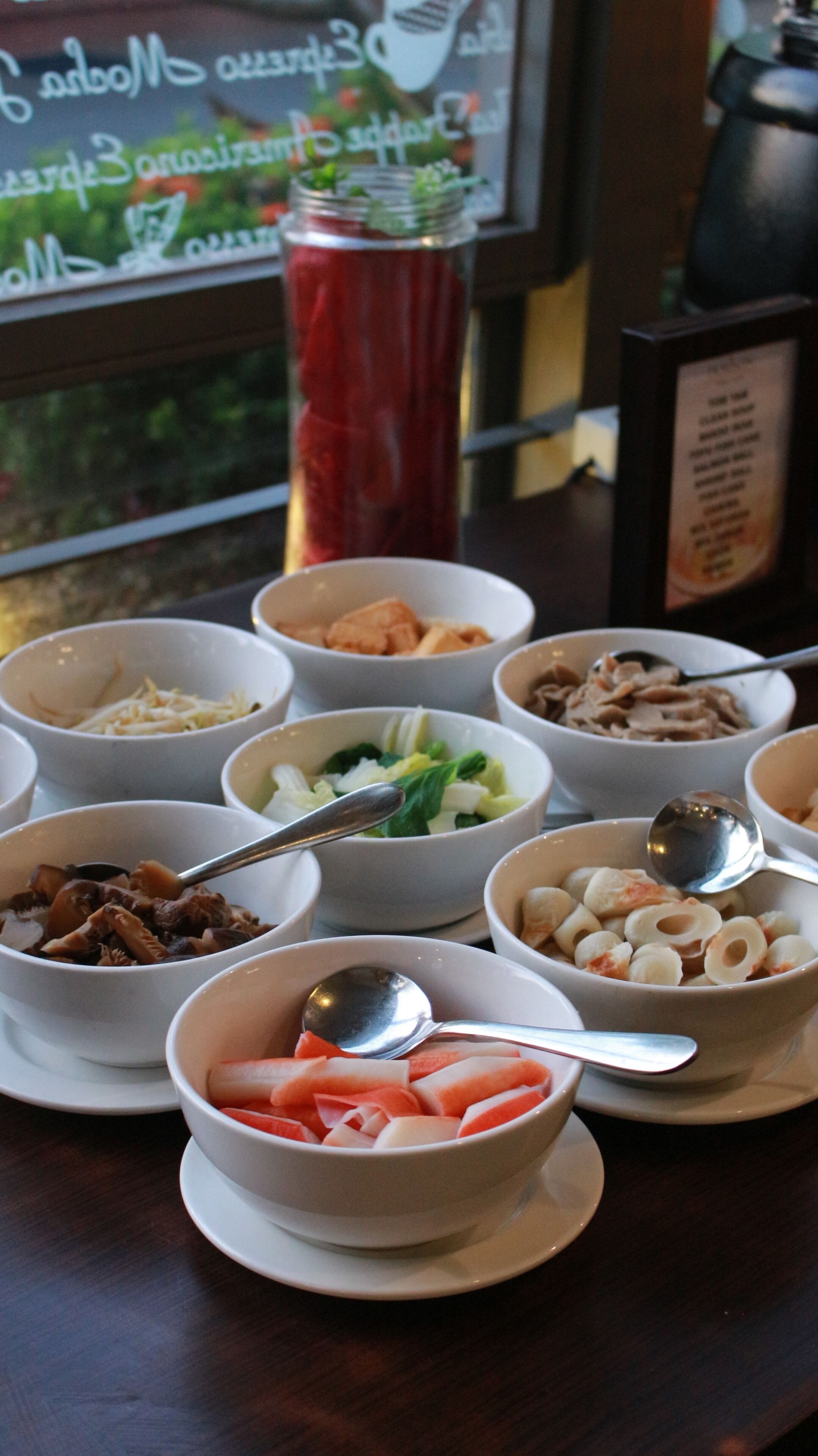 A table with various bowls of food, spoons, and a red container.