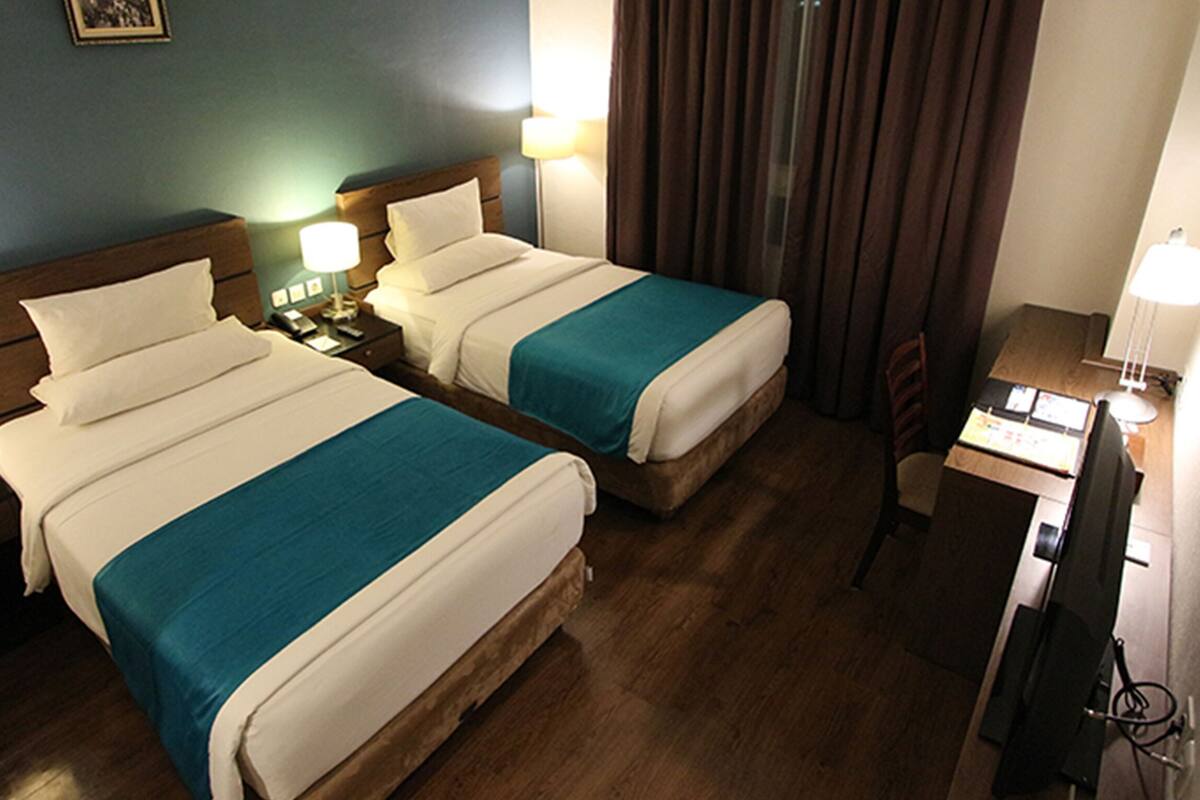 A hotel room with two beds, wooden headboards, bedside tables with lamps, a desk with a computer, and a window with curtains.
