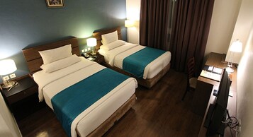 Habitación de hotel con dos camas, cabeceros de madera, mesitas de noche con lámparas, un escritorio con ordenador y una ventana con cortinas.
