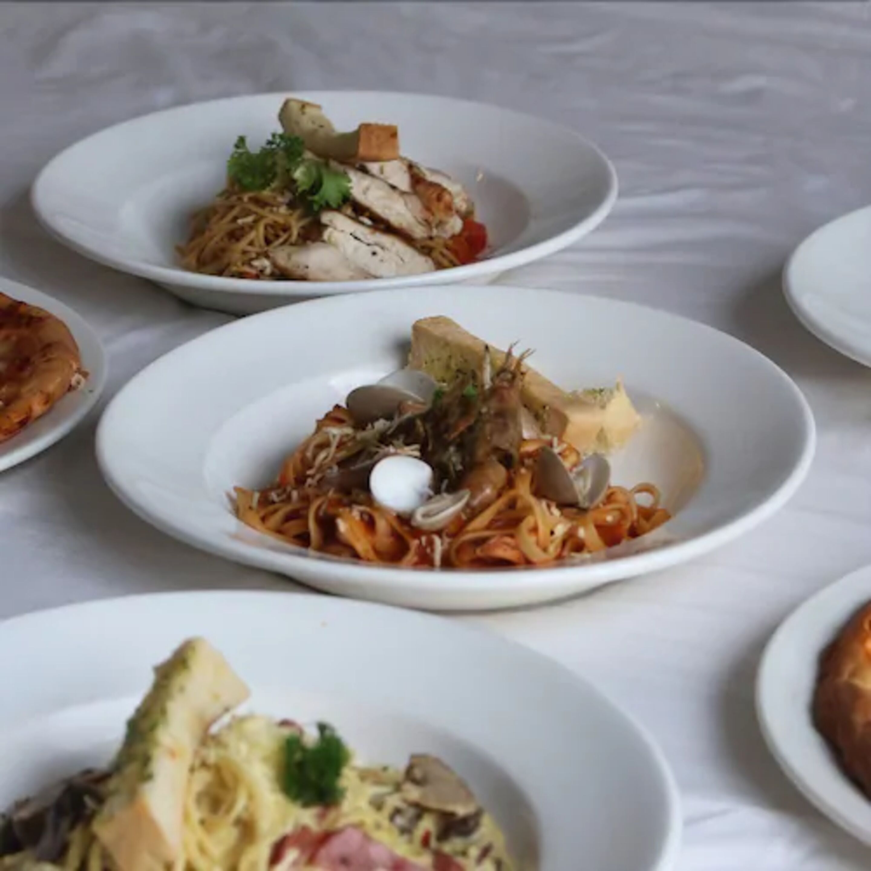 Flera vita tallrikar med olika rätter, bland annat pasta, skaldjur och grönsaker, uppradade på ett bord.