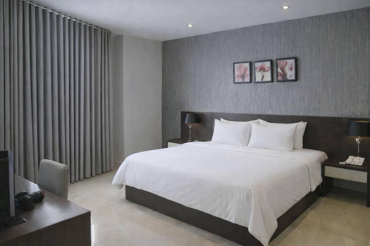 Una habitación de hotel moderna con una cama grande, dos mesitas de noche con lámparas, un escritorio con computadora y cámara, y un ventanal con persianas verticales.
