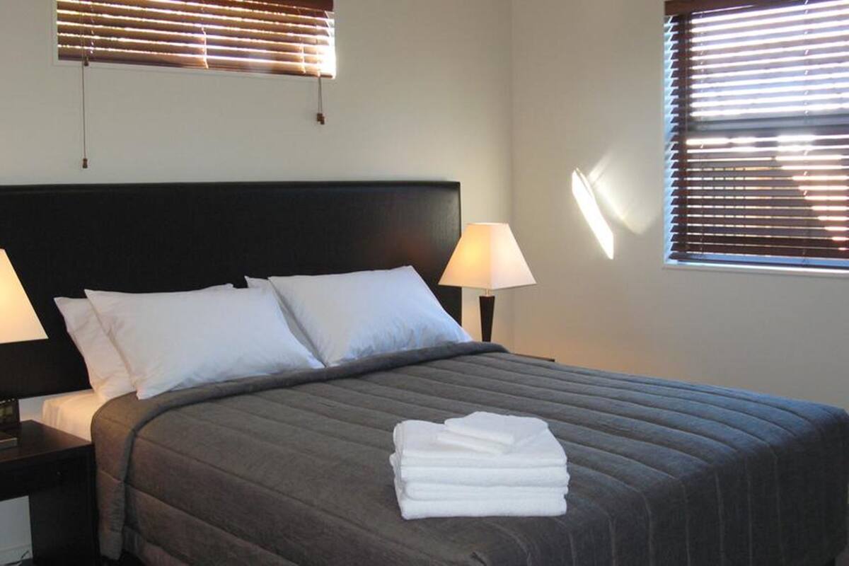 A neatly made bed with a dark headboard, two bedside lamps, and a window with blinds.