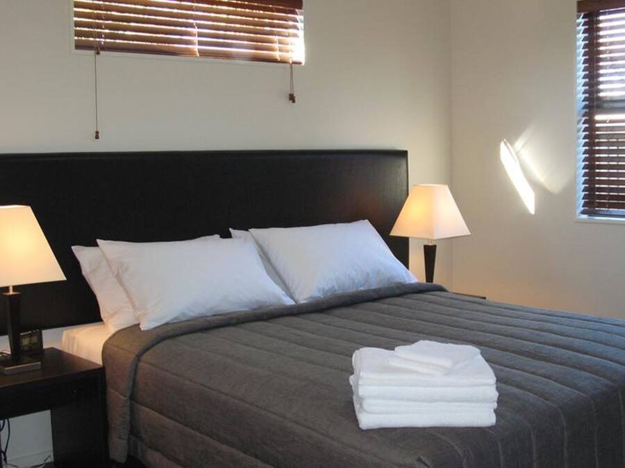 A neatly made bed with a dark headboard, two bedside lamps, and a window with blinds.
