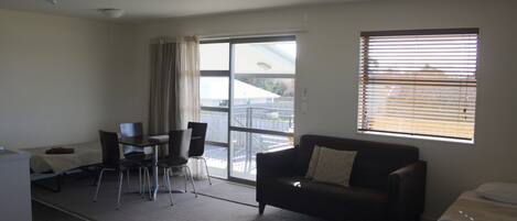 Habitación de hotel con cama, sofá, escritorio con silla, mesa de centro y una ventana grande con persianas.
